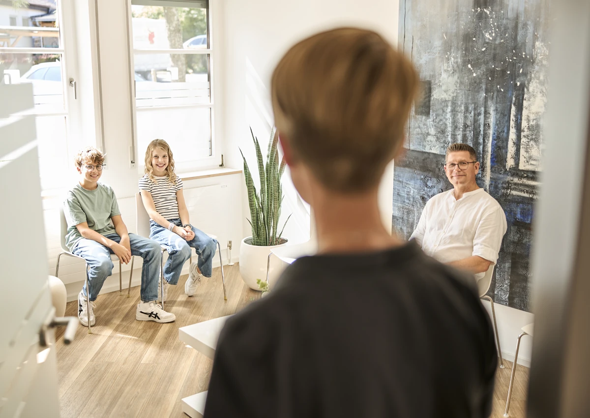Personen im Wartezimmer einer Zahnarztpraxis, darunter zwei Kinder und ein Erwachsener.