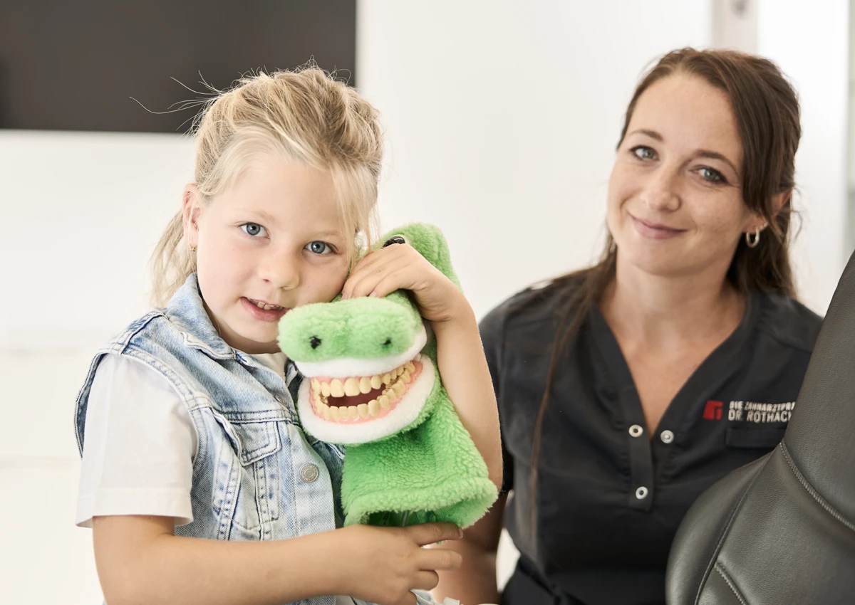 Kind hält ein Plüschtier-Krokodil im Arm. Das Kind und eine Assistentin lächeln in die Kamera.