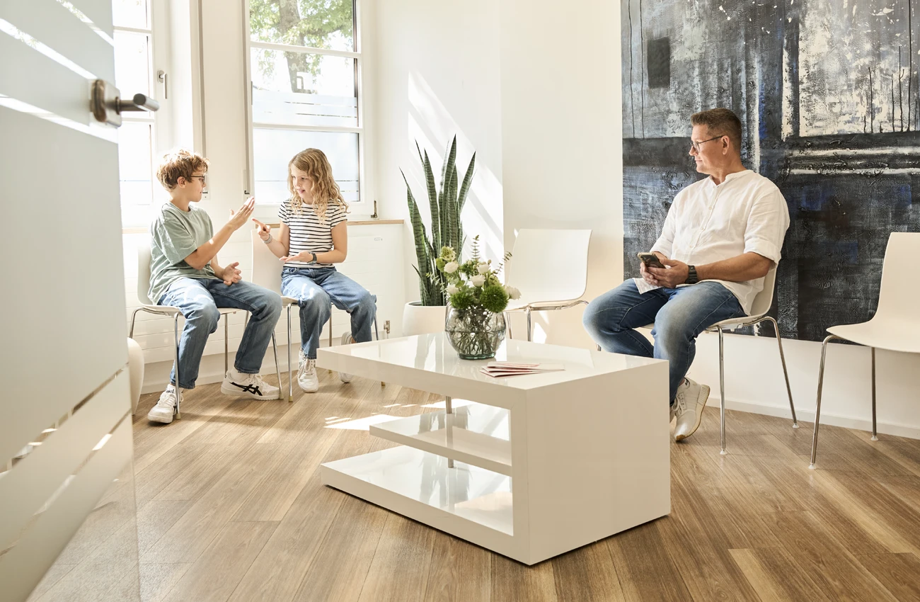 Wartezimmer bei Dr. Rothacher: Zwei Kinder spielen, ein Mann liest auf seinem Handy. Moderne und helle Einrichtung.
