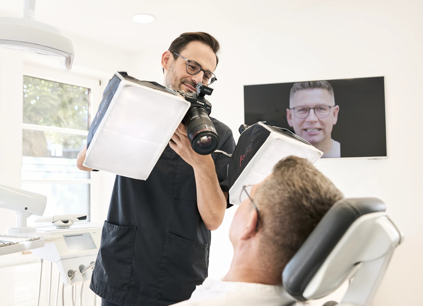 Zahnarzt fotografiert Patienten im Behandlungsstuhl, Bildschirm im Hintergrund zeigt lächelndes Porträt.